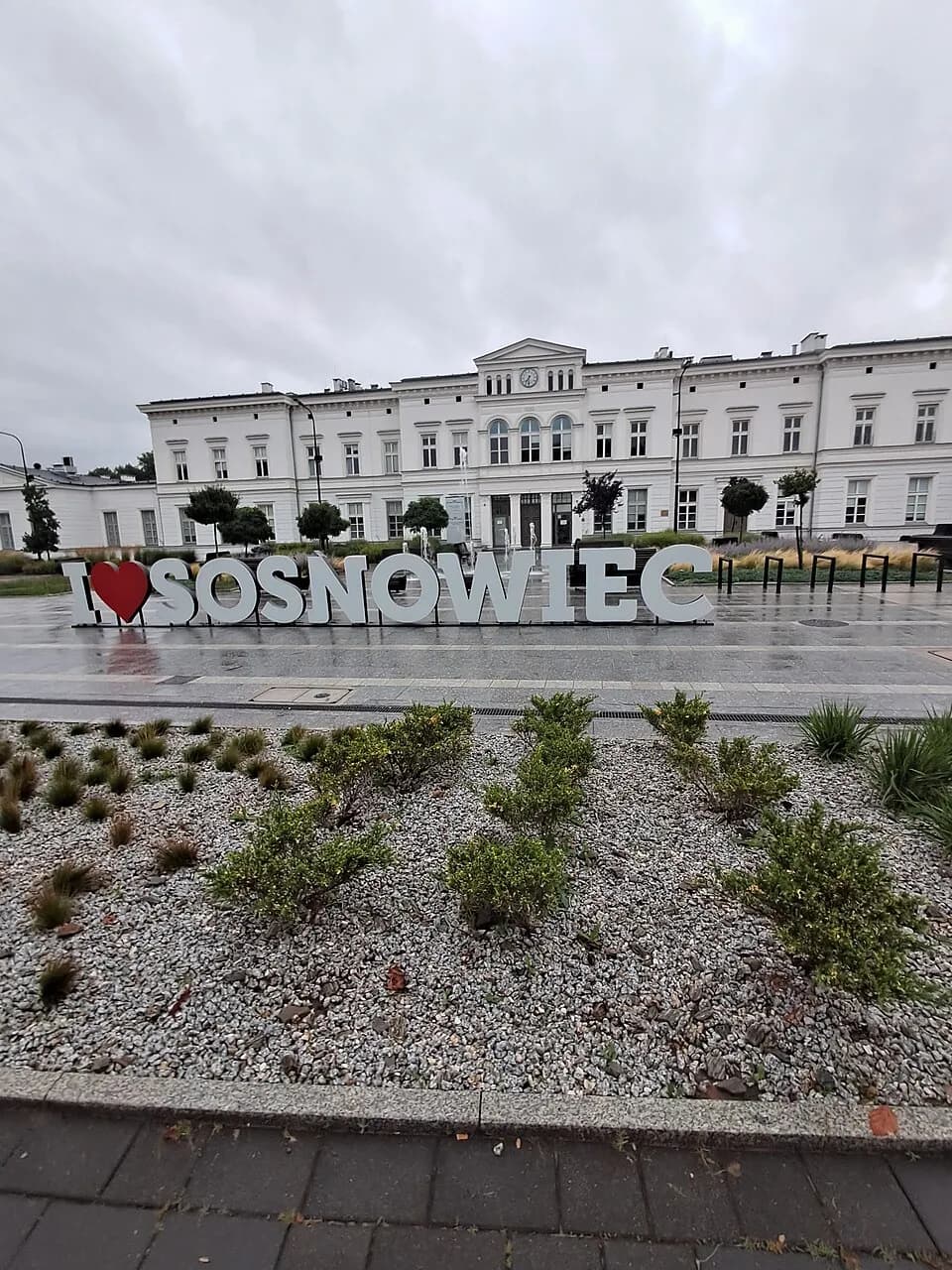 A large sign displaying "I heart SOSNOWIEC" in front of a grand, white classical building with a clock tower under an overcast sky.