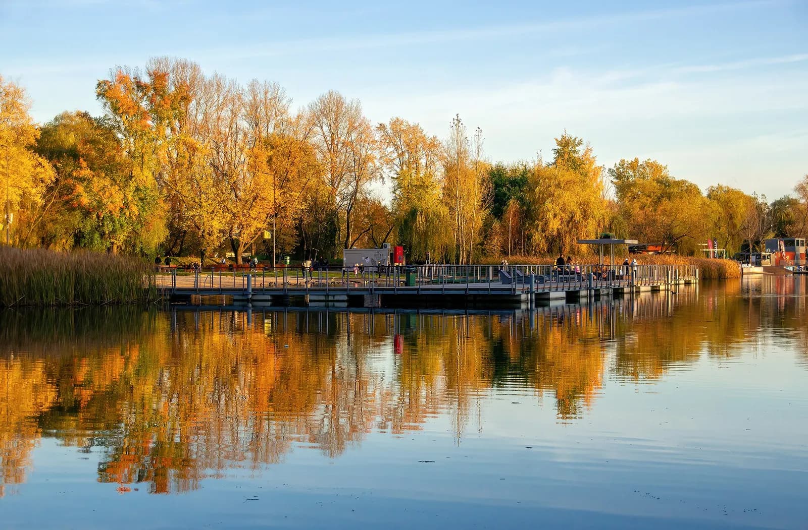 A long pier extends into a calm lake, surrounded by trees with vibrant autumn foliage reflected in the water under a clear sky.