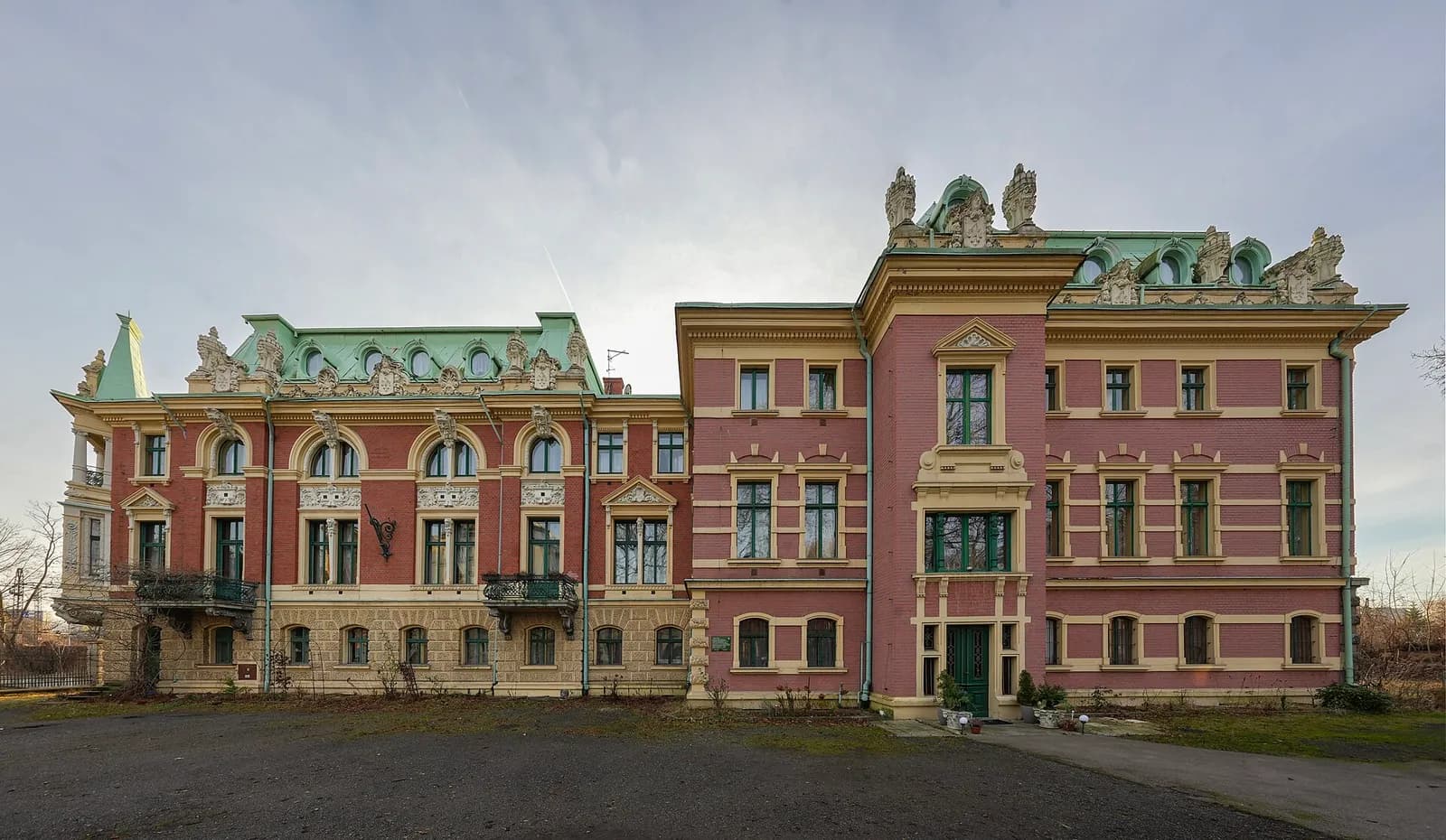 A grand, salmon-colored manor with ornate green roofs, multiple windows, and a dark paved area in front under a cloudy sky.