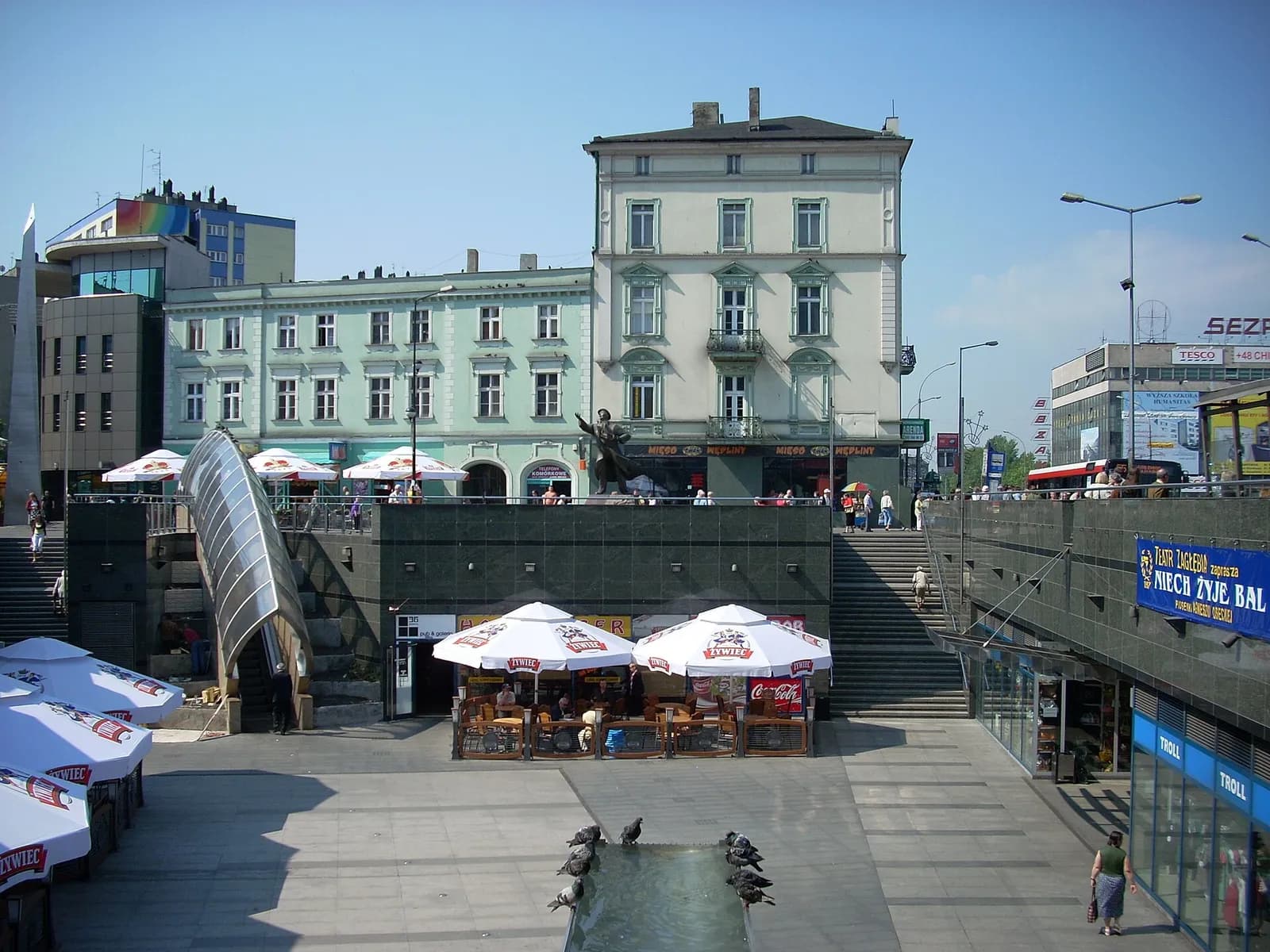 A bustling town square features a sculpture, multiple cafes with umbrellas, and a fountain, surrounded by historic buildings and modern architecture under a clear blue sky.